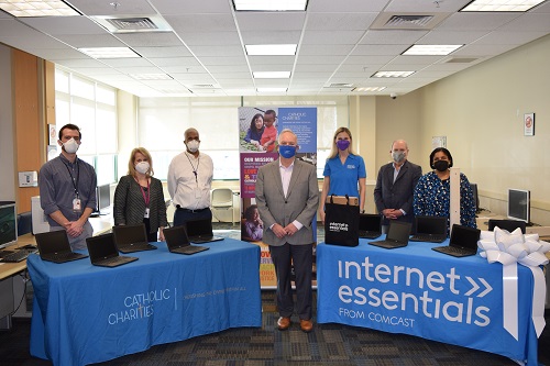 Catholic Charities colleagues Kevin Creamer, Sue DeSantis, Lee Martin, Bill MCarthy and Amy Collier with representatives from Comcast stand behind two tables with laptop computers open on them.